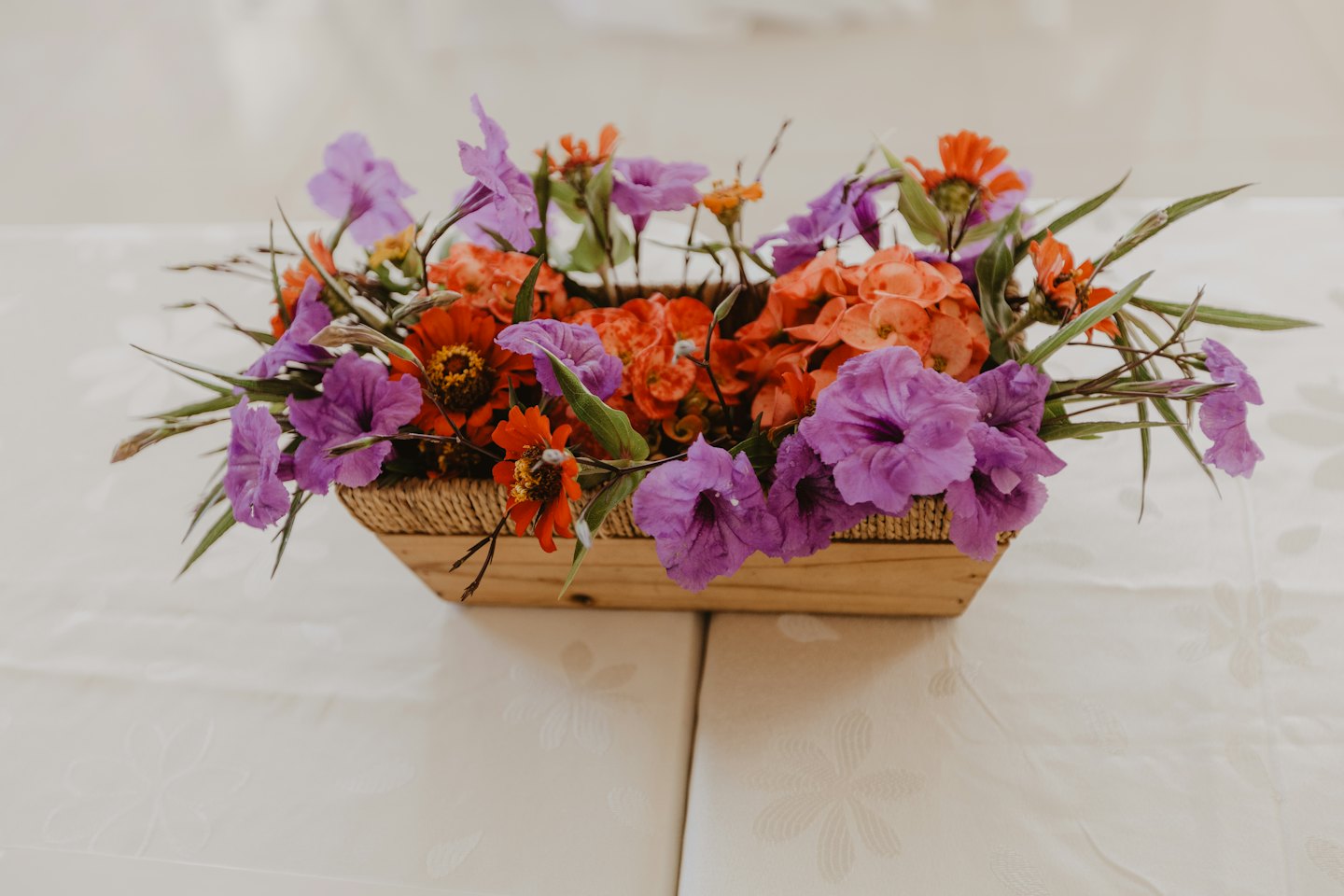 Purple and orange flowers in a wooden basket on a tablecloth.